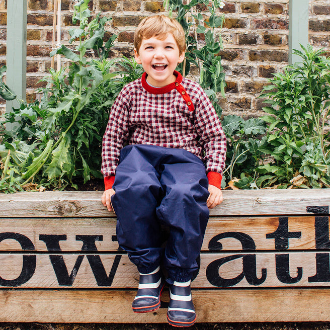 A young boy sits smiling in a garden, wearing Originals Waterproof Trousers Navy, a chequered red and white top and stripy wellington boots.